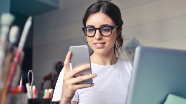 A young woman looks at her smart phone whilst sat in front of her laptop