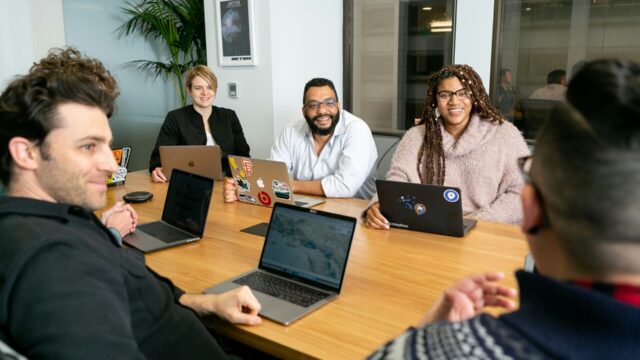 four people all on laptops, two men and two women, listen to person talking in a board meeting